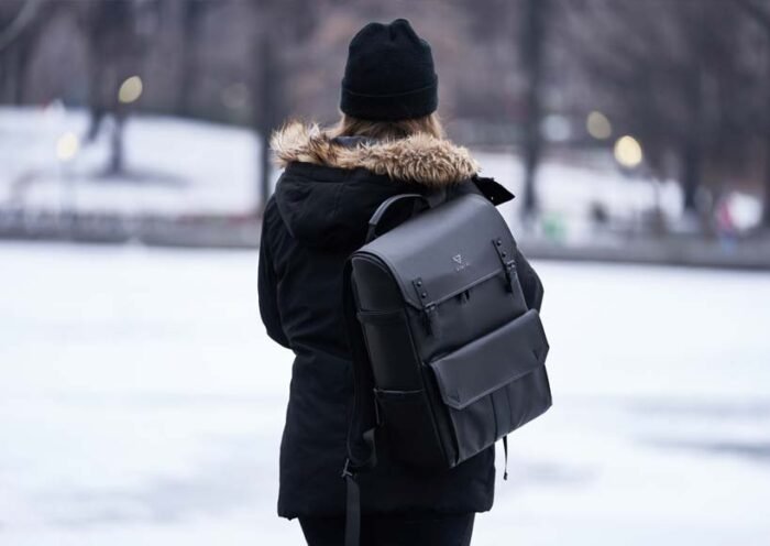 Person wearing a black winter coat with a fur trim hood and a black backpack standing outdoors in a snowy park in back view