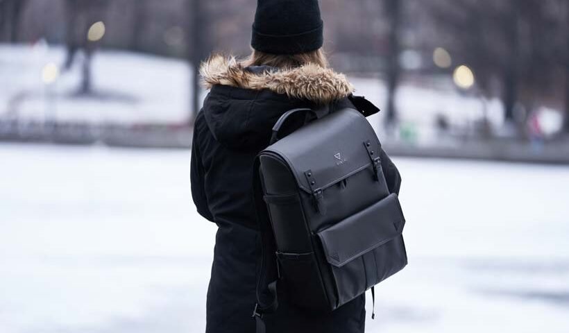 Person wearing a black winter coat with a fur-trim hood and a black backpack standing outdoors in a snowy park in back view.