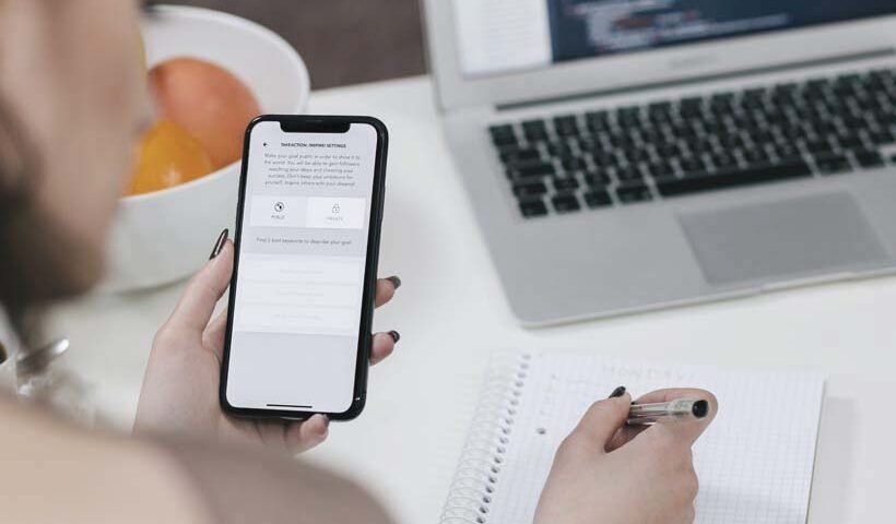Person using a mobile app on a phone while taking notes in a spiral notebook, with a laptop open nearby.
