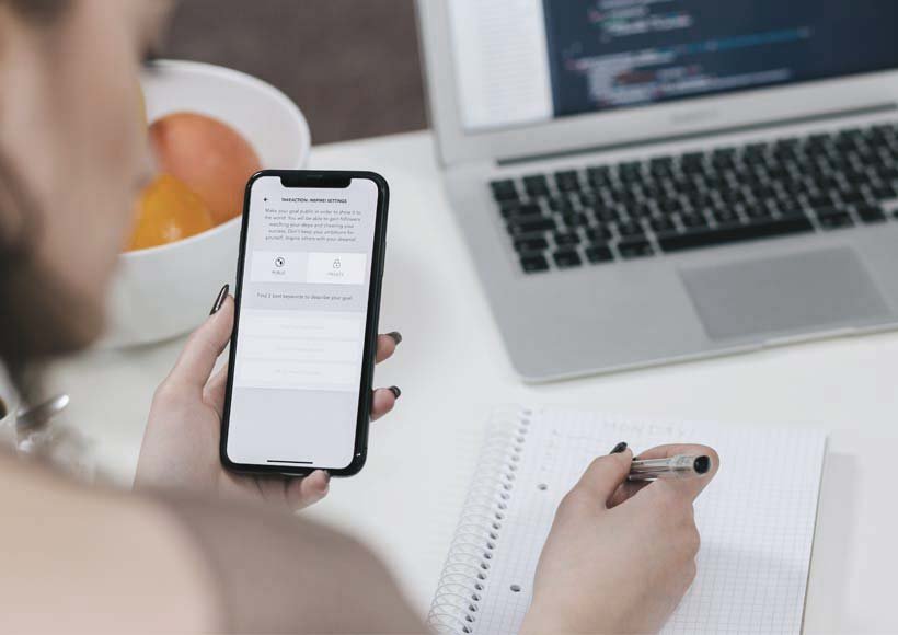 Person using a mobile app on a phone while taking notes in a spiral notebook with a laptop open nearby