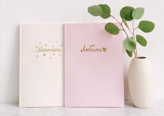Two pastel notebooks cream and pink with gold cyrillic text leaning on a white surface beside a white vase with eucalyptus leaves