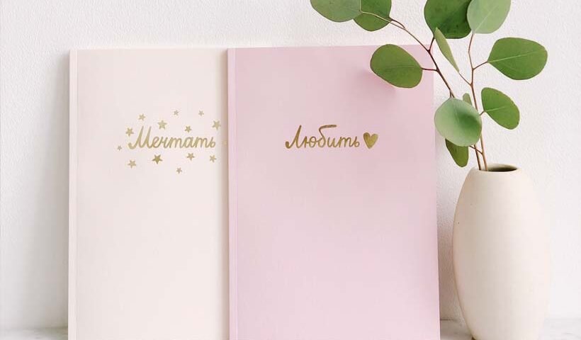 Two pastel notebooks (cream and pink) with gold Cyrillic text, leaning on a white surface beside a white vase with eucalyptus leaves.