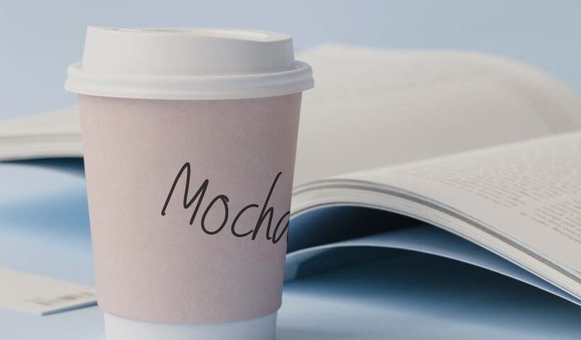To-go cup labeled Mocha on a desk beside an open magazine.