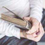 Person sitting with a light sweater holding a worn hardcover book in their lap