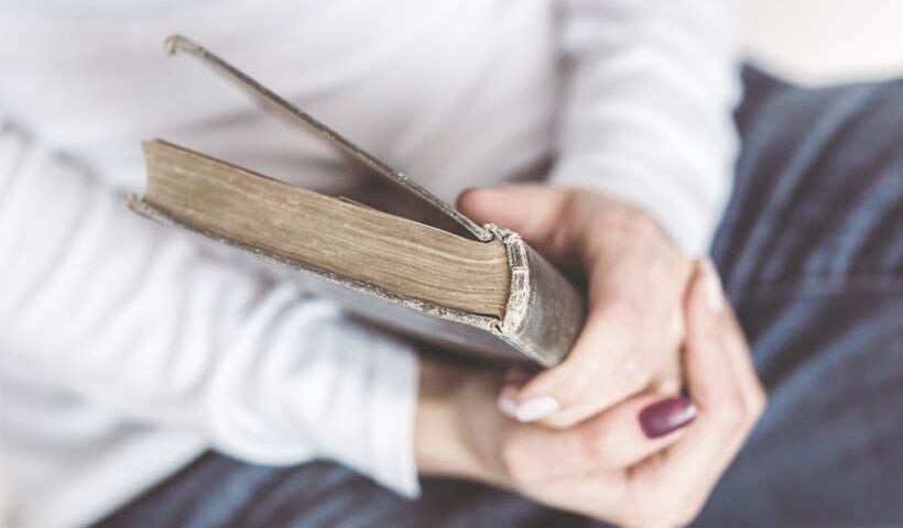 Person sitting with a light sweater, holding a worn hardcover book in their lap.