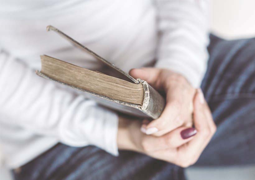 Person sitting with a light sweater holding a worn hardcover book in their lap