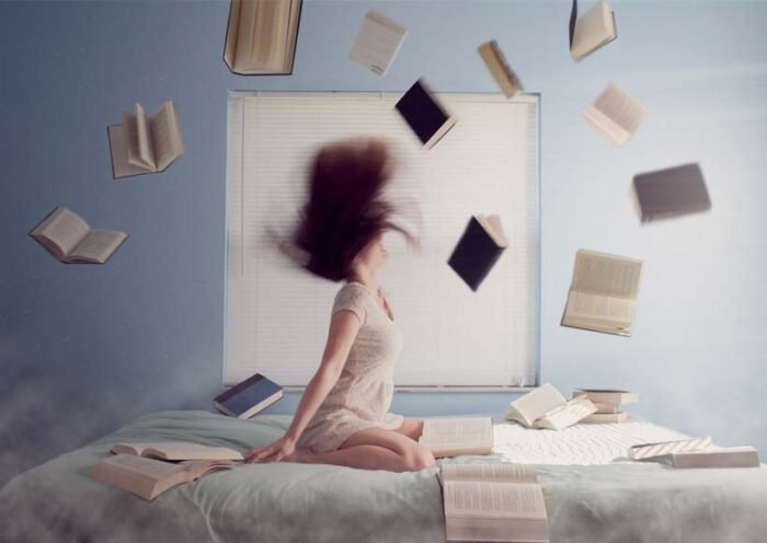Young woman sitting on a bed while books fly around her in a chaotic study scene