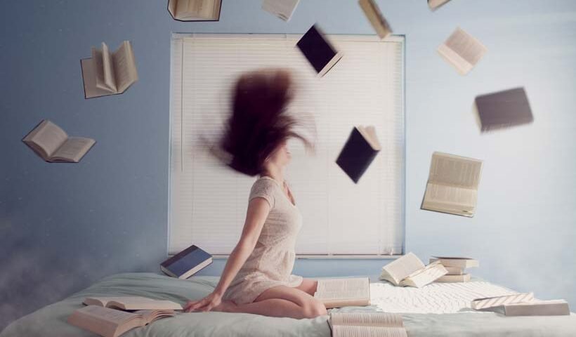 Young woman sitting on a bed while books fly around her in a chaotic study scene.