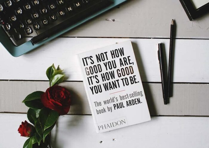 Desk scene with a white book titled its not how good you are beside a keyboard a rose and writing pens on a light wooden surface