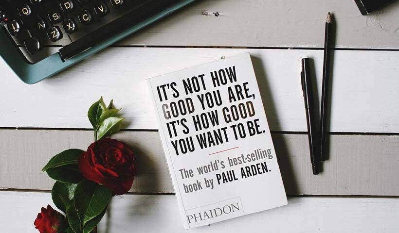 Desk scene with a white book titled 'It’s Not How Good You Are…' beside a keyboard, a rose, and writing pens on a light wooden surface.