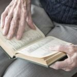 Elderly hands turning the pages of an open hardcover book resting on the lap