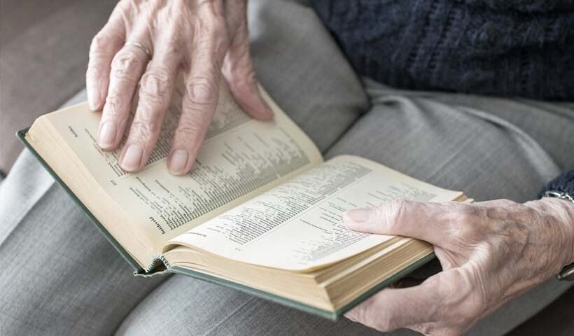 Elderly hands turning the pages of an open hardcover book resting on the lap.
