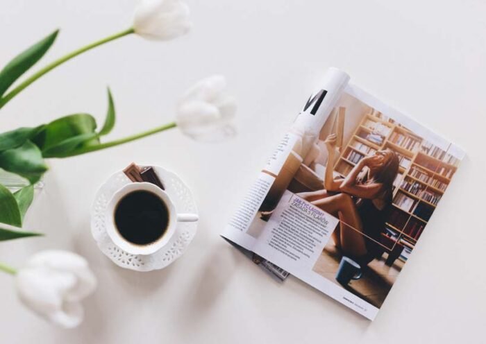 A cup of black coffee on a white saucer beside an open magazine featuring a woman in a library with white tulips in the background