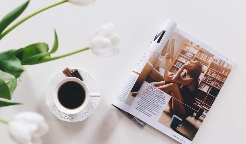 A cup of black coffee on a white saucer beside an open magazine featuring a woman in a library, with white tulips in the background.