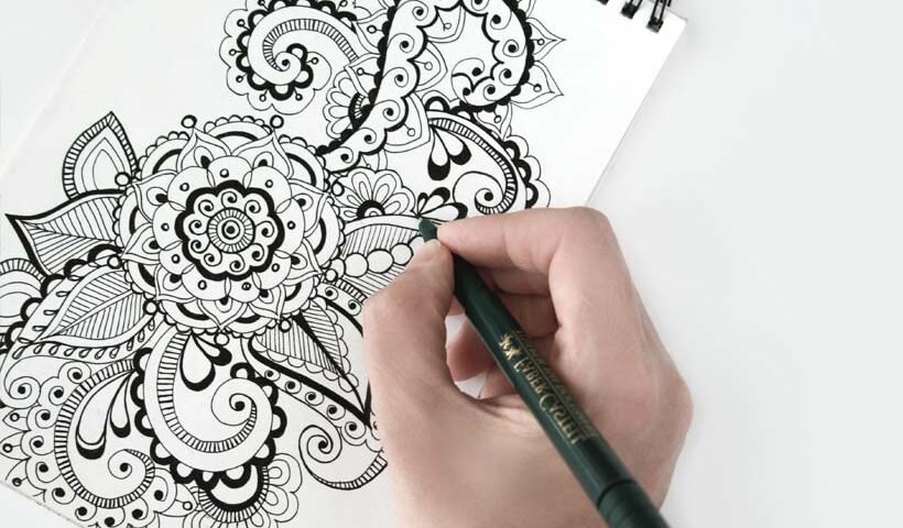Close-up of a hand drawing an intricate black-and-white mandala pattern in a spiral notebook with a pen poised to draw.