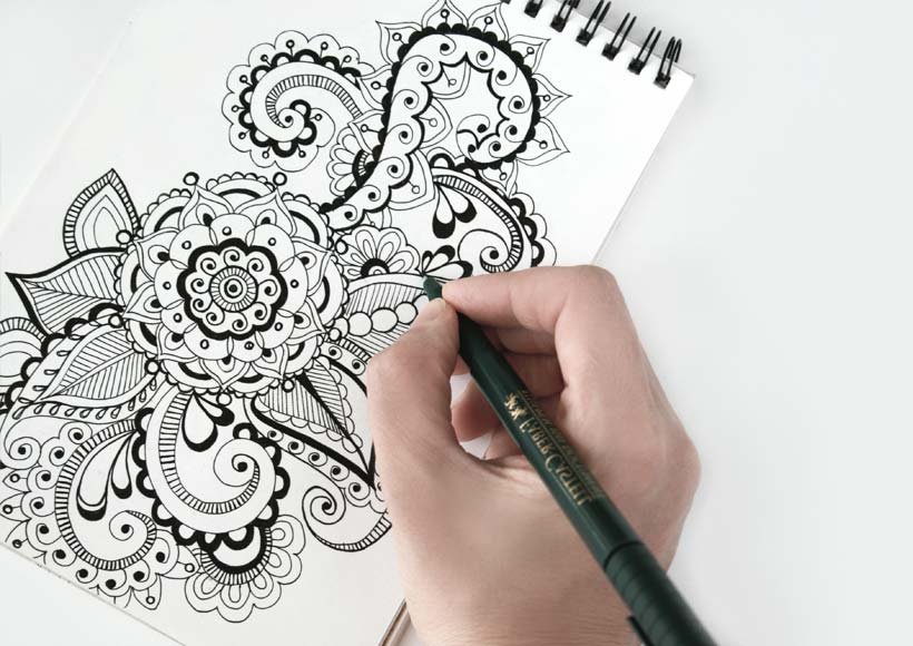 Close up of a hand drawing an intricate black and white mandala pattern in a spiral notebook with a pen poised to draw