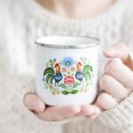 Hands in a cream knit sweater hold a white enamel mug with a colorful floral folk art design