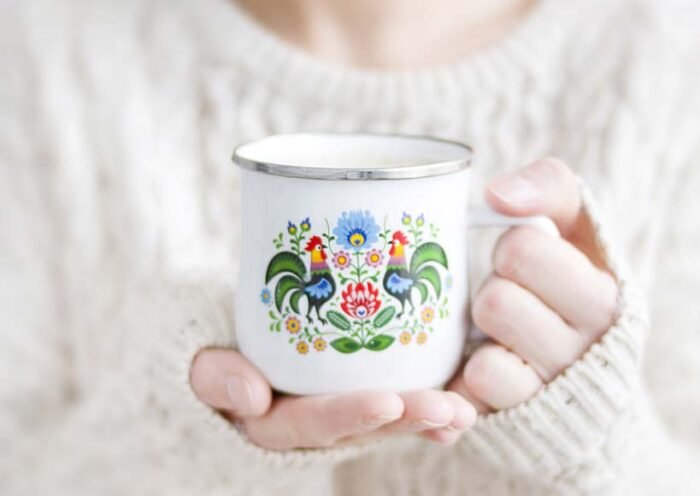 Hands in a cream knit sweater hold a white enamel mug with a colorful floral folk art design