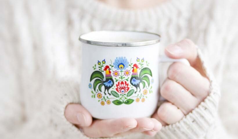 Hands in a cream knit sweater hold a white enamel mug with a colorful floral folk-art design.