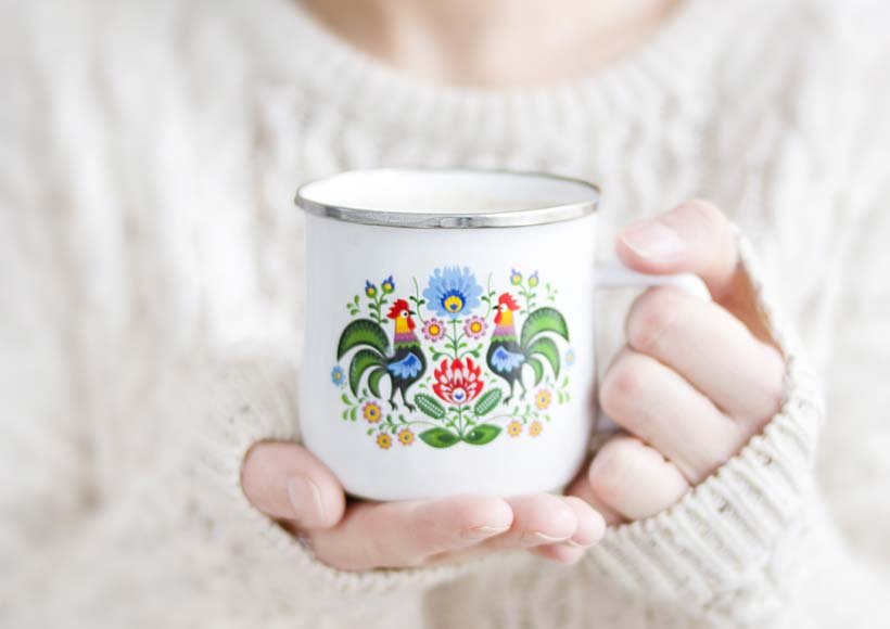 Hands in a cream knit sweater hold a white enamel mug with a colorful floral folk art design