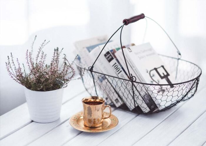 Copper cup and saucer sit on a white wooden table beside a potted plant with a wire basket of newspapers in the background