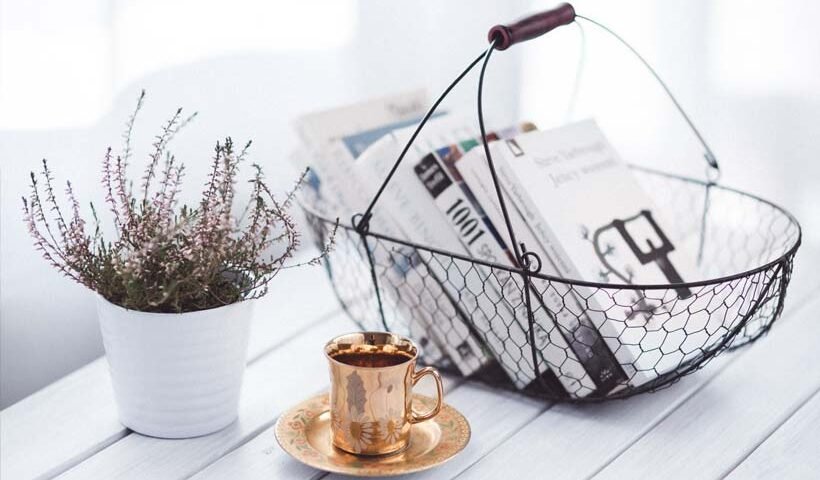 Copper cup and saucer sit on a white wooden table beside a potted plant, with a wire basket of newspapers in the background.