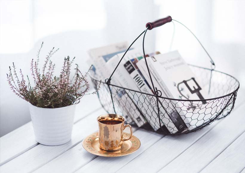 Copper cup and saucer sit on a white wooden table beside a potted plant with a wire basket of newspapers in the background