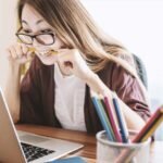 Woman wearing glasses bites a pencil while looking at a laptop screen at a desk cluttered with colorful pencils in a pencil holder