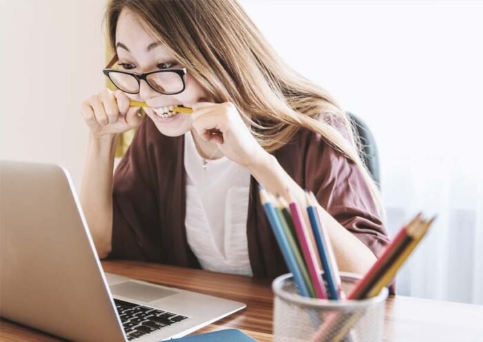 Woman wearing glasses bites a pencil while looking at a laptop screen at a desk cluttered with colorful pencils in a pencil holder