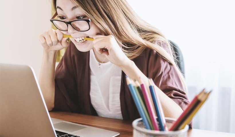 Woman wearing glasses bites a pencil while looking at a laptop screen at a desk cluttered with colorful pencils in a pencil holder.