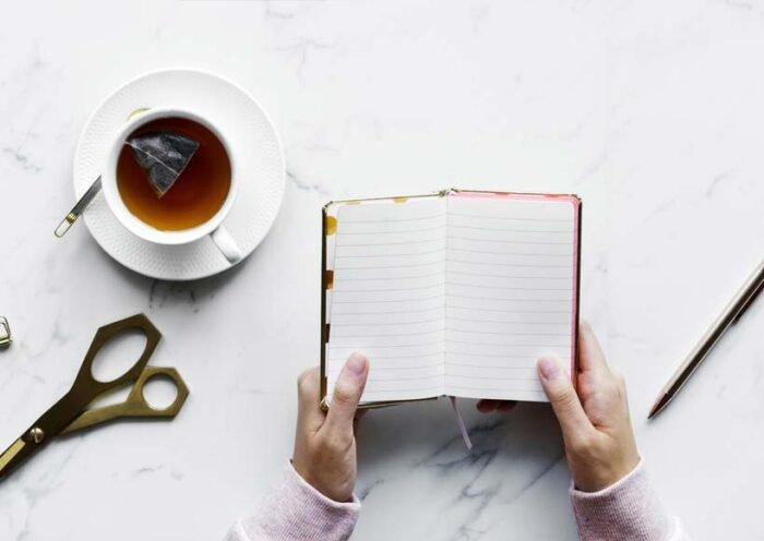 Top down view of a person holding an open notebook with lined pages a cup of tea with a teabag scissors and a pencil on a marble desk