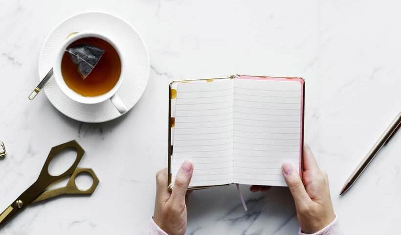 Top-down view of a person holding an open notebook with lined pages, a cup of tea with a teabag, scissors, and a pencil on a marble desk