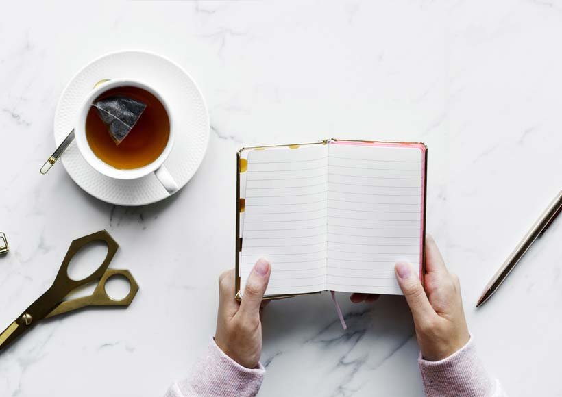 Top down view of a person holding an open notebook with lined pages a cup of tea with a teabag scissors and a pencil on a marble desk