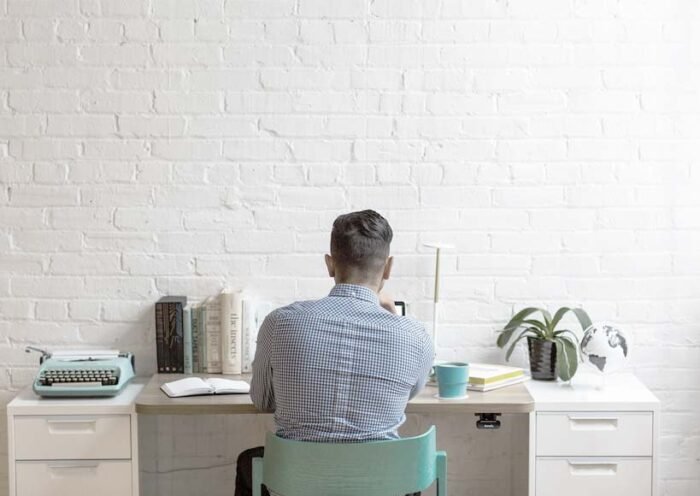 Back view of a person in a checkered shirt sitting at a white desk in a bright home office typing or focusing on work with books a typewriter plant and mug nearby