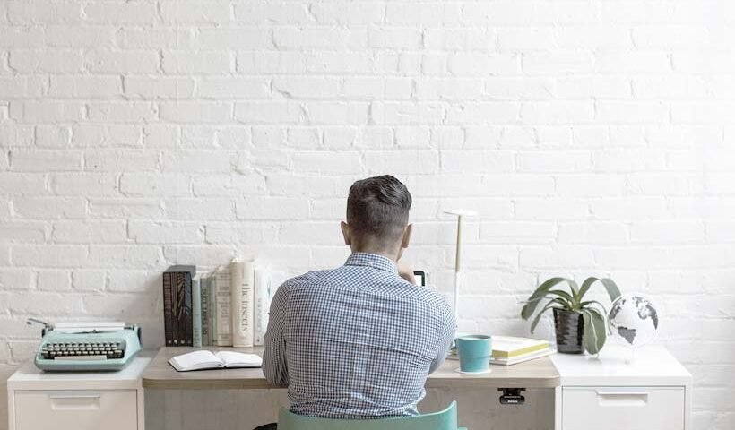 Back view of a person in a checkered shirt sitting at a white desk in a bright home office, typing or focusing on work with books, a typewriter, plant, and mug nearby