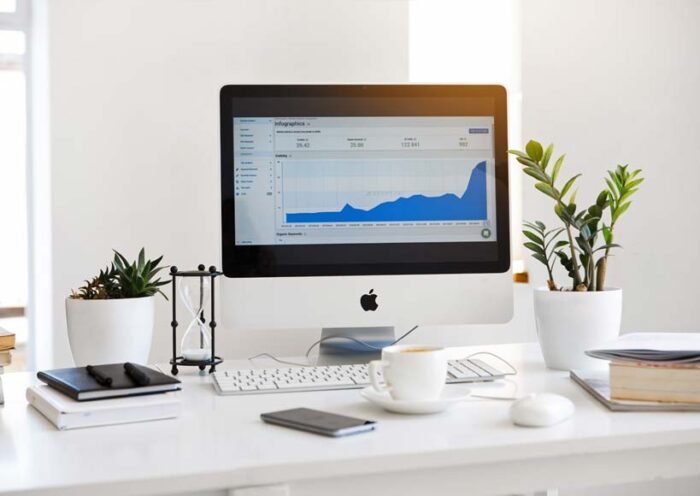 Desk workspace with an imac displaying a blue analytics chart flanked by potted plants and office supplies on a white desk