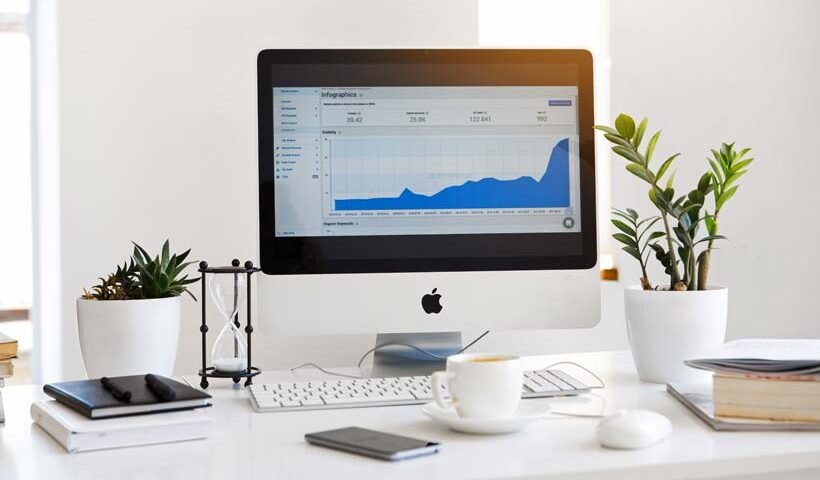 Desk workspace with an iMac displaying a blue analytics chart, flanked by potted plants and office supplies on a white desk.