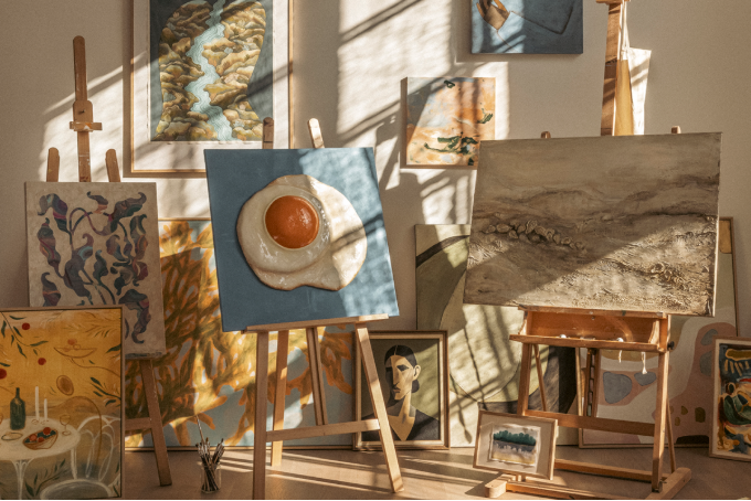 Sunlit art studio with multiple easels displaying various paintings including a blue canvas with a fried egg a landscape and abstract pieces